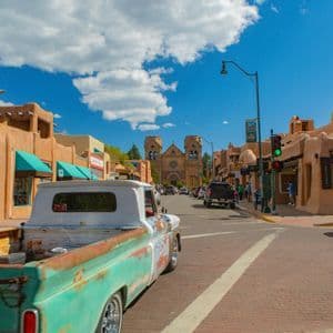Un pickup vintage turchese su una strada di mattoni fiancheggiata da edifici in stile adobe, con una grande cattedrale visibile alla fine della strada sotto un cielo blu.