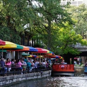 La gente mangia in un ristorante sul fiume sotto ombrelloni colorati mentre due battelli turistici navigano lungo il canale alberato.