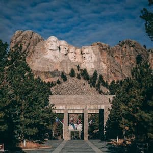 Il Monte Rushmore National Memorial visto dal vialetto d'ingresso in pietra, fiancheggiato da pini sotto un cielo azzurro.