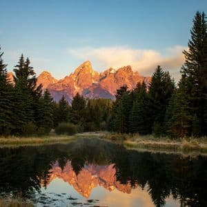 Cime montuose frastagliate illuminate dal sole si riflettono in un lago immobile, con una fitta foresta sempreverde in primo piano.