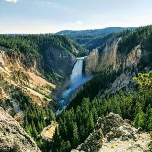 Una grande cascata precipita in un fiume all'interno di un vasto canyon densamente boscoso di pini sotto un cielo azzurro.