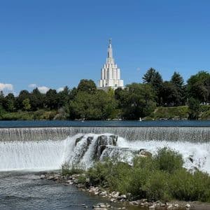 Un'ampia cascata scende lungo un fiume, con un tempio bianco a più livelli che si erge sopra gli alberi sulla sponda opposta sotto un cielo azzurro.