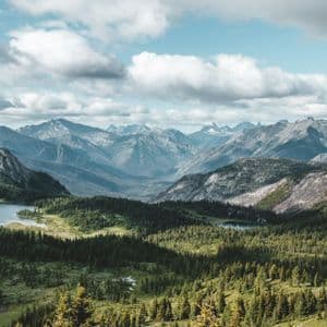 Una vasta catena montuosa con cime innevate si affaccia su una valle lussureggiante con foreste e piccoli laghi sotto un cielo azzurro nuvoloso.