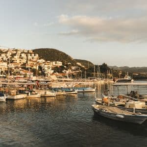 Numerose piccole imbarcazioni sono ormeggiate in un porto soleggiato di fronte a una cittadina costiera su un fianco di collina verdeggiante.