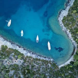 Vista aerea di barche a vela ancorate in una caletta cristallina e turchese, adiacente a una costa rocciosa e boscosa.