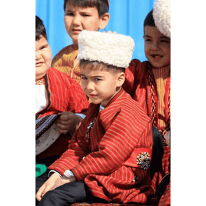 Un bambino, con una giacca tradizionale a righe rosse e un cappello di lana bianco, siede in un gruppo di bambini, guardando la fotocamera.