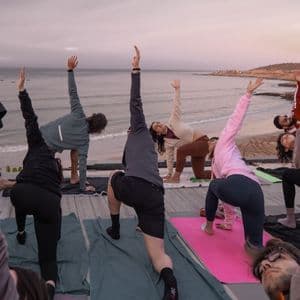 Un gruppo WeRoad pratica yoga su un pontile di legno con vista su spiaggia e oceano all'alba.