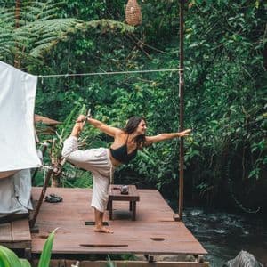 Una donna pratica una posizione yoga su un ponte di legno accanto a un fiume che scorre in una giungla verde e fitta.