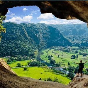 A person with arms outstretched stands at the mouth of a cave, looking out over a lush green valley with mountains and a river.