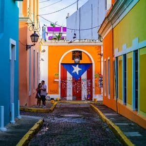 A narrow cobblestone alley flanked by colorful buildings, with a large door at the end painted with the flag of Puerto Rico.