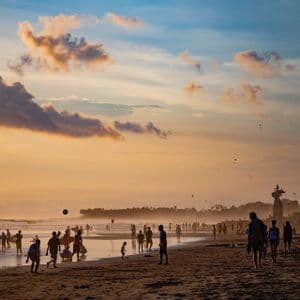 Sagome di persone su una spiaggia affollata al tramonto, che giocano e passeggiano in riva al mare sotto un cielo dorato.