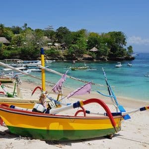 Barche tradizionali colorate riposano su una spiaggia di sabbia bianca accanto a una baia calma e turchese con una costa verde sullo sfondo.