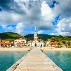 Une jetée en bois s'avance sur des eaux turquoise vers une ville côtière avec une église, adossée à des collines verdoyantes sous un ciel nuageux.