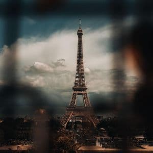 The Eiffel Tower is viewed from a distance against a dramatic, cloudy sky, with blurry elements framing the shot.