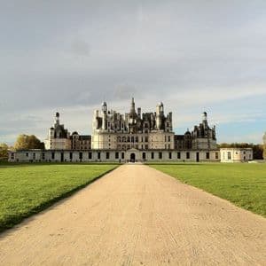 A long, unpaved path leads across a vast green lawn to a large, ornate white castle with many towers under a partly cloudy sky.
