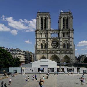 The facade of Notre Dame Cathedral on a sunny day, with people walking in the large plaza in front of the reconstruction site.