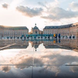 A grand historical building and cloudy sky are reflected in the wet surface of a large city plaza at sunset.
