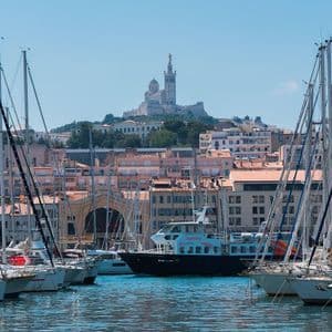 A bustling harbor filled with sailboats and a tour boat, with a city and a prominent basilica on a hill in the background.