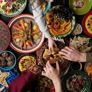 Una vista superior de manos que se extienden hacia varios platos durante un festín festivo de Eid, incluyendo tayín, frutas y pan plano.