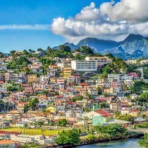 Case e palazzi colorati raggruppati su una verdeggiante collina, con montagne rigogliose sullo sfondo sotto un cielo parzialmente nuvoloso.