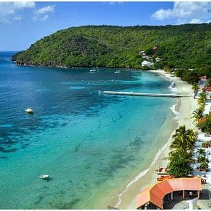 Vue aérienne d'une ville côtière avec une plage de sable, une jetée s'avançant dans une eau turquoise et une colline verdoyante.
