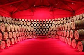 Dans une cave illuminée par une lumière rouge intense, des dizaines de tonneaux en bois sont empilés le long d'une grande paroi incurvée.