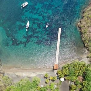 Veduta aerea di un lungo molo che si estende in una cala dalle acque turchesi, con diverse barche ancorate vicino a una costa boscosa.