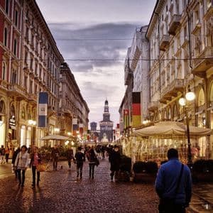 People walk down a cobblestone street lined with illuminated shops and flags at dusk, with a historic castle visible in the distance.
