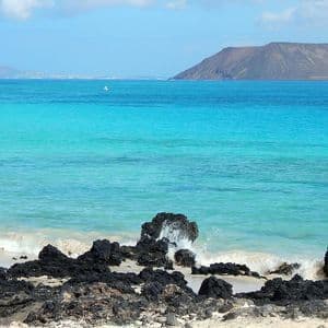 Las olas rompen contra rocas volcánicas negras en una playa de arena, con agua turquesa y una gran isla al fondo.