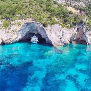 Un viaggio di gruppo WeRoad su una barca turistica che entra in una grande grotta marina alla base di una scogliera alberata, circondata da acqua turchese e cristallina.