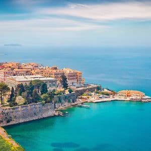 Una vista dall'alto di una città costiera con tetti in terracotta e mura fortificate che domina una baia turchese.