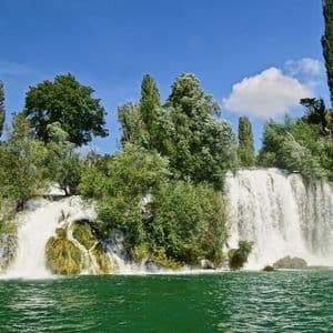 Una cascata ampia si getta in un fiume verde, circondata da alberi rigogliosi sotto un cielo azzurro limpido.