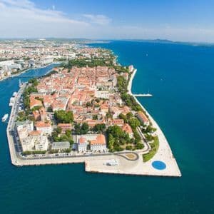 Vista aerea di una città storica su una penisola con edifici dai tetti arancioni, circondata da acque calme e di un blu profondo sotto un cielo sereno.