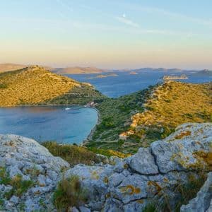 Una vista mozzafiato da una collina rocciosa che domina una tranquilla baia blu, circondata da colline e isole illuminate dal sole sotto un cielo limpido.