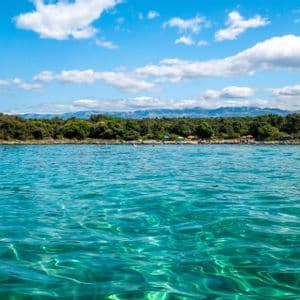 Acque turchesi cristalline con una riva alberata e montagne distanti visibili sotto un cielo azzurro parzialmente nuvoloso.