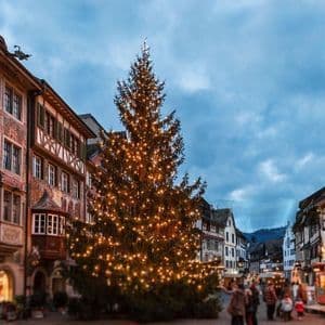 Un grande albero di Natale illuminato si erge in una storica piazza europea con persone che si godono un mercatino di Natale al tramonto.