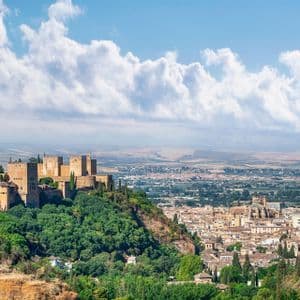 Una antigua fortaleza de piedra se asienta sobre una exuberante colina verde, dominando una extensa ciudad bajo un cielo azul con nubes esponjosas.