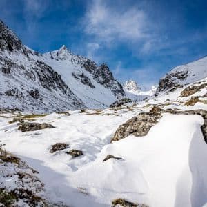 Un amplio valle montañoso cubierto de nieve con grandes rocas y picos escarpados bajo un cielo azul brillante con nubes tenues.