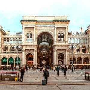People walk across a large, paved square in front of the ornate facade and grand archway of a historic building.