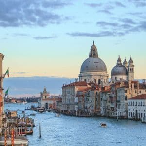 Boats and a gondola travel along a wide canal flanked by historic buildings, with a large domed basilica in the distance under a cloudy sky.