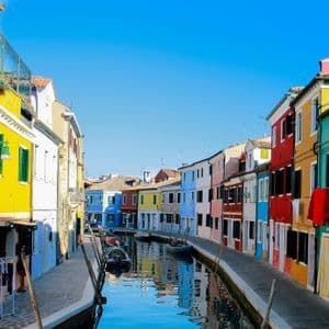 Brightly colored houses line a narrow water canal with several moored boats under a clear blue sky.