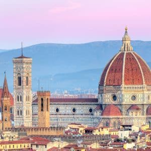 A large cathedral with a red-tiled dome and a tall bell tower overlooking a cityscape at dusk, with mountains in the distance.