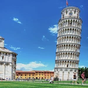 The Leaning Tower of Pisa and a cathedral stand on a green lawn with tourists walking by under a bright blue sky.