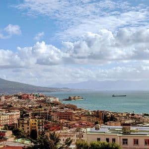 A panoramic view of a colorful coastal city, a bay, and a large volcano under a partly cloudy sky.