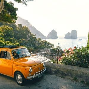 An orange vintage car with an open sunroof parked on a scenic coastal road overlooking a sunlit sea with large rock formations.