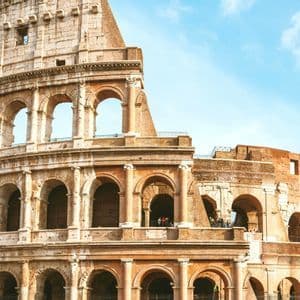A sunlit, partial view of the ancient Roman Colosseum's tiered stone arches against a bright blue sky with light clouds.