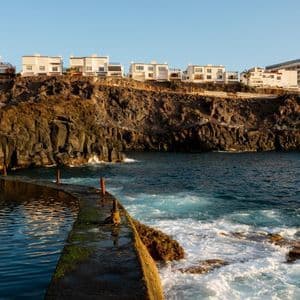 Edifici bianchi sulla costa si ergono su scogliere rocciose, affacciati su una piscina naturale e sull'oceano, con onde che si infrangono contro una passerella in pietra.