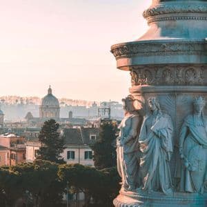 A close-up of carved marble statues on a monument, with a hazy city skyline featuring a large dome in the background.