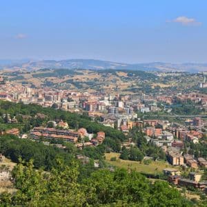 Un'ampia e panoramica vista di una città adagiata in una valle, circondata da verdi colline ondulate sotto un cielo azzurro e limpido.
