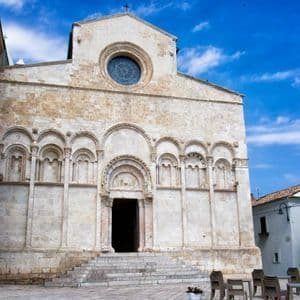 La facciata in pietra di una chiesa romanica con portali ad arco e un grande rosone, situata in una piazza cittadina.